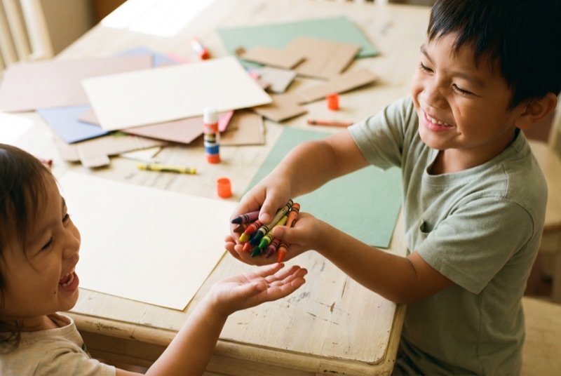 A boy handing a fistful of crayons to a smaller child across a craft table.