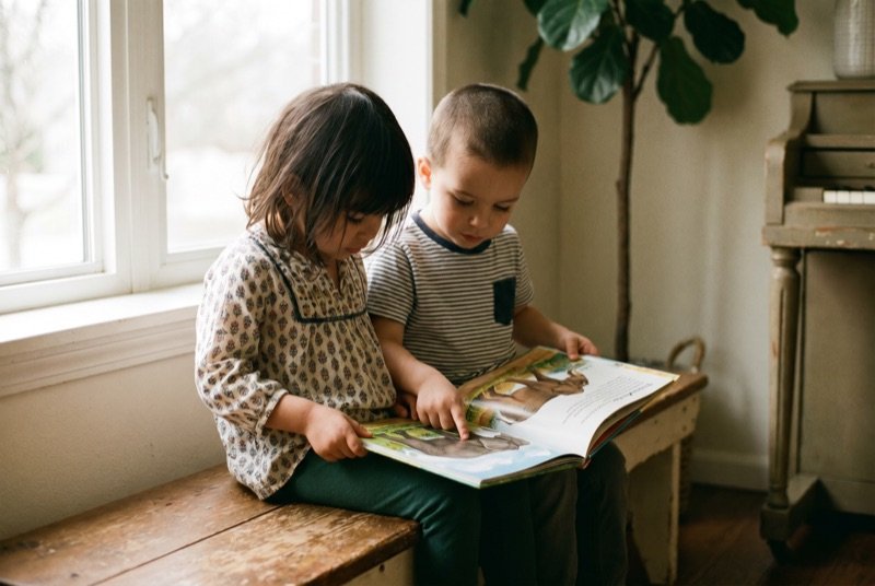 Two small children sitting on a bench, poring over a large picture Bible together.